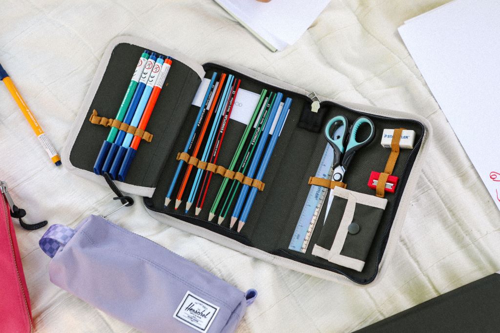 Penalhus med indhold, farveblyanter, sakse og viskelæder på hvidt underlag