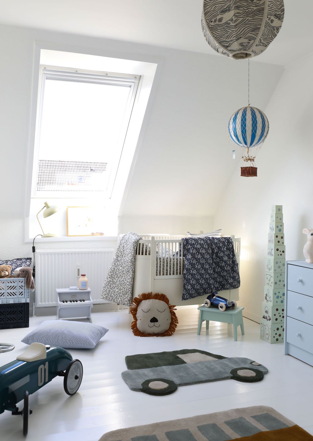 Børneværelse med tremmeseng, lyse vægge og dekorationer, herunder luftballon og løvepude.